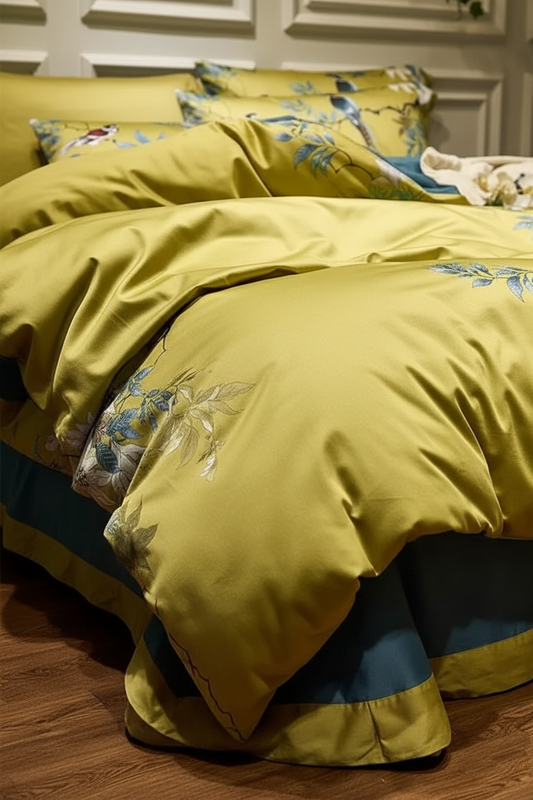 A close-up of a bed dressed in a yellow-green satin bedding set with blue and white floral embroidery, set against a warm wood-paneled wall and wooden floor.