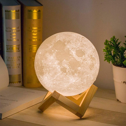 A realistic full moon-shaped LED desk lamp illuminated, placed on a wooden stand, with books and a potted plant in the background.