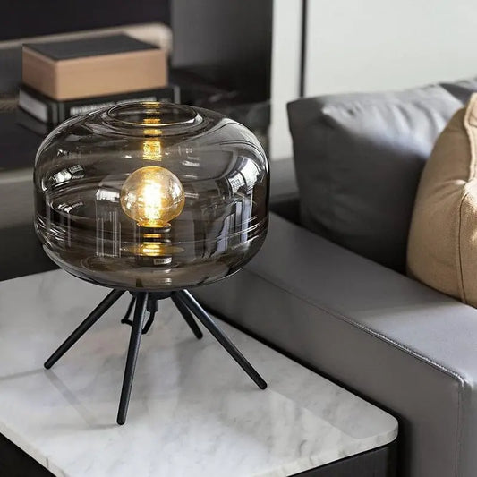 Smoky glass table lamp with a black metal base on a marble surface in a living room setting.